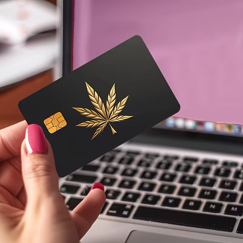 Hand holding a black card with a gold cannabis leaf design in front of a laptop.